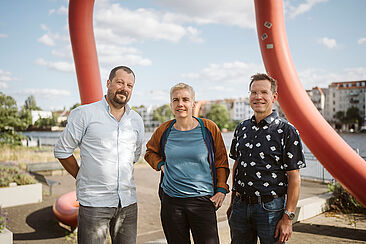 Drei Personen stehen vor einer großen roten Skulptur im Freien mit urbaner Kulisse im Hintergrund. Es handelt sich um die Preisträger*innen des Preises für gute Lehre 2025 an der HTW Berlin: Stephen Mortimer, Juliane Siegeris und Thomas Hücker.