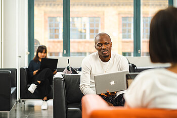 A student works on a laptop in an open learning space while others study in the background.