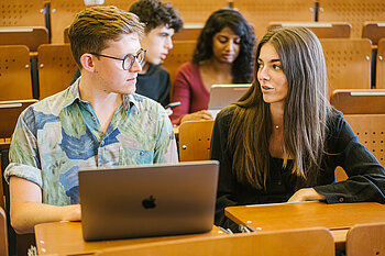 Two students sit in a lecture hall and discuss their work on a laptop.