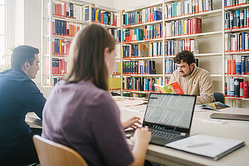 Students study together in the library surrounded by well-stocked bookshelves.